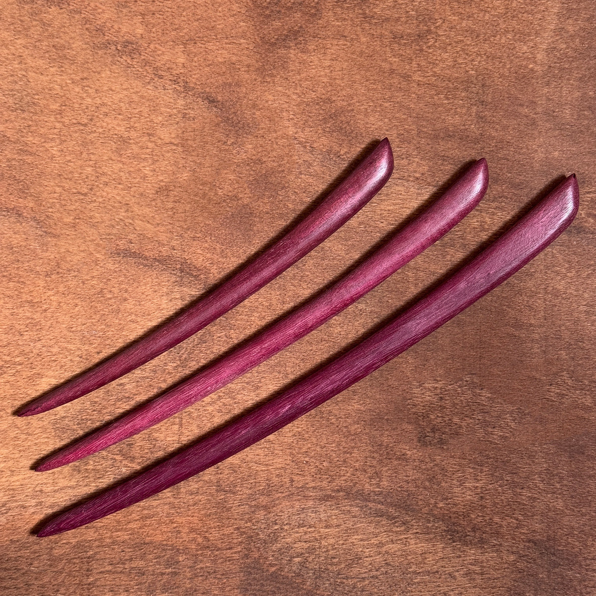 A clear, well-lit view of three beautiful purpleheart wood hair sticks resting on a wooden surface.
