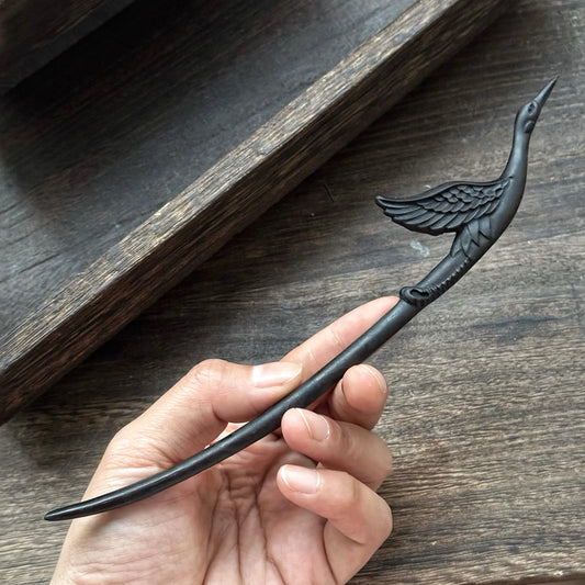 A hand holding the detailed carving of the crane on the 18cm black ebony wood hair stick to show scale.