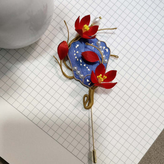 Ronghua handmade plum blossom fan brooch with blue fan base, gold trim, three red silk plum flowers with yellow stamens, pearl accents, and gold wire branches on a long stick pin, displayed on white grid paper