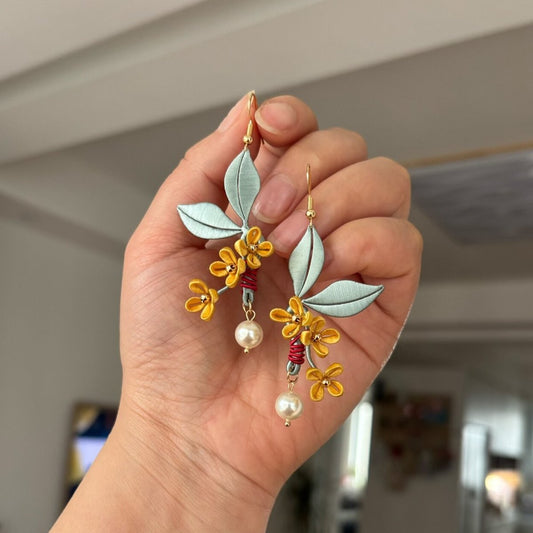 Chanhua handmade osmanthus drop earrings with golden yellow flower clusters, soft sage green leaves, red wire stem accent, and round pearl drop, held in hand against an indoor background