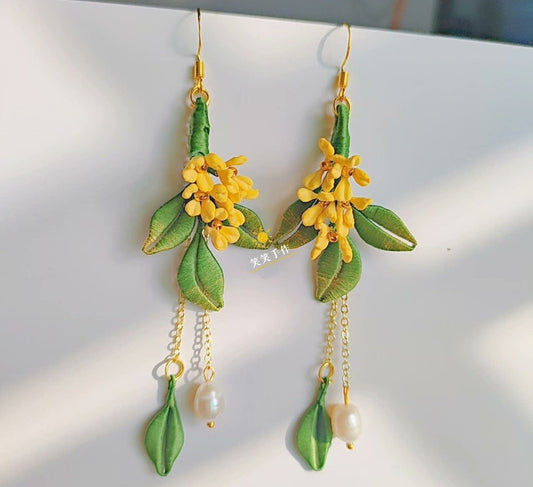 Pair of artisanal Chanhua earrings featuring detailed yellow osmanthus flowers, green silk leaves, and dangling freshwater pearls, photographed against a bright backdrop.