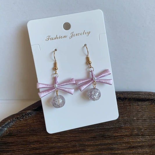 A pair of handmade Chanhua earrings with a light pink ribbon bow and a clear, shimmering beaded drop.