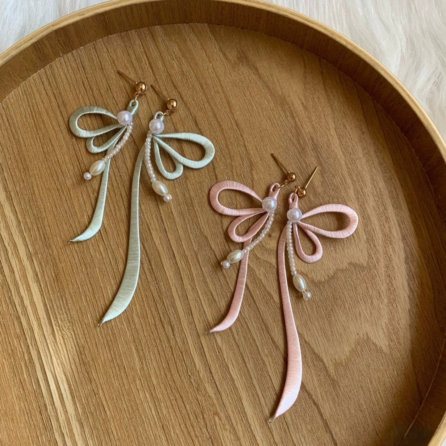 A pair of handmade light green and light pink Chanhua ribbon earrings with freshwater pearls, lying on a wooden tray.