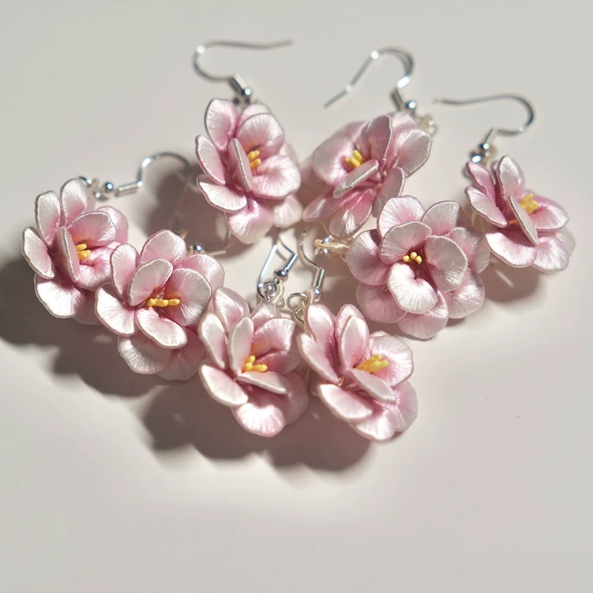 A collection of multiple pairs of handmade pink camellia flower earrings with 925 sterling silver hooks, laid out on a white surface.