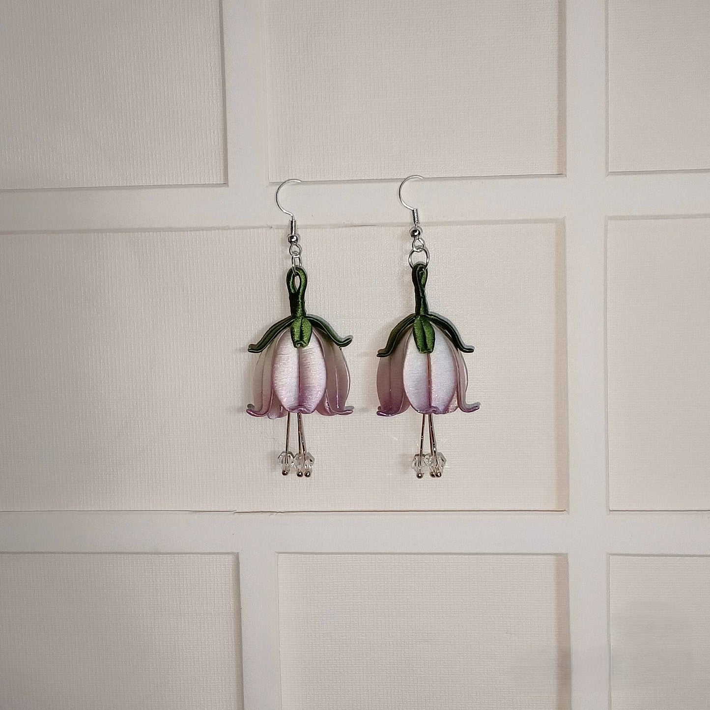 A pair of handmade Chanhua flower dangle earrings on a white textured background. The earrings feature light purple petals and a green stem, with small crystal beads at the bottom.