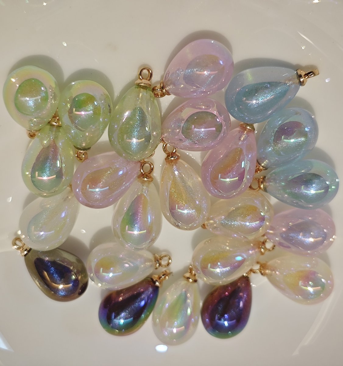 Assorted colors of iridescent galaxy waterdrop charms on a white plate.