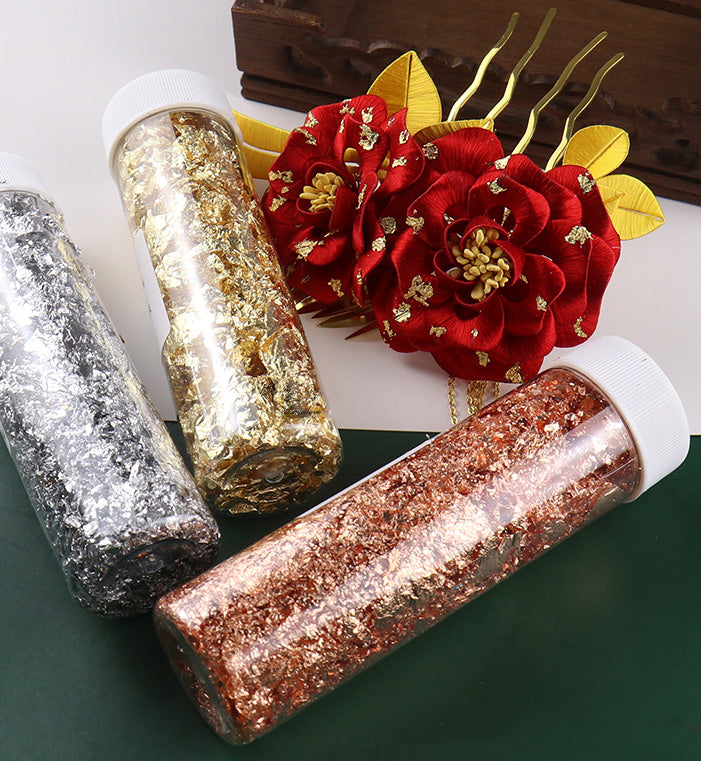 Three jars of metallic craft flakes in gold, silver, and rose gold, displayed next to a traditional red floral hair accessory.
