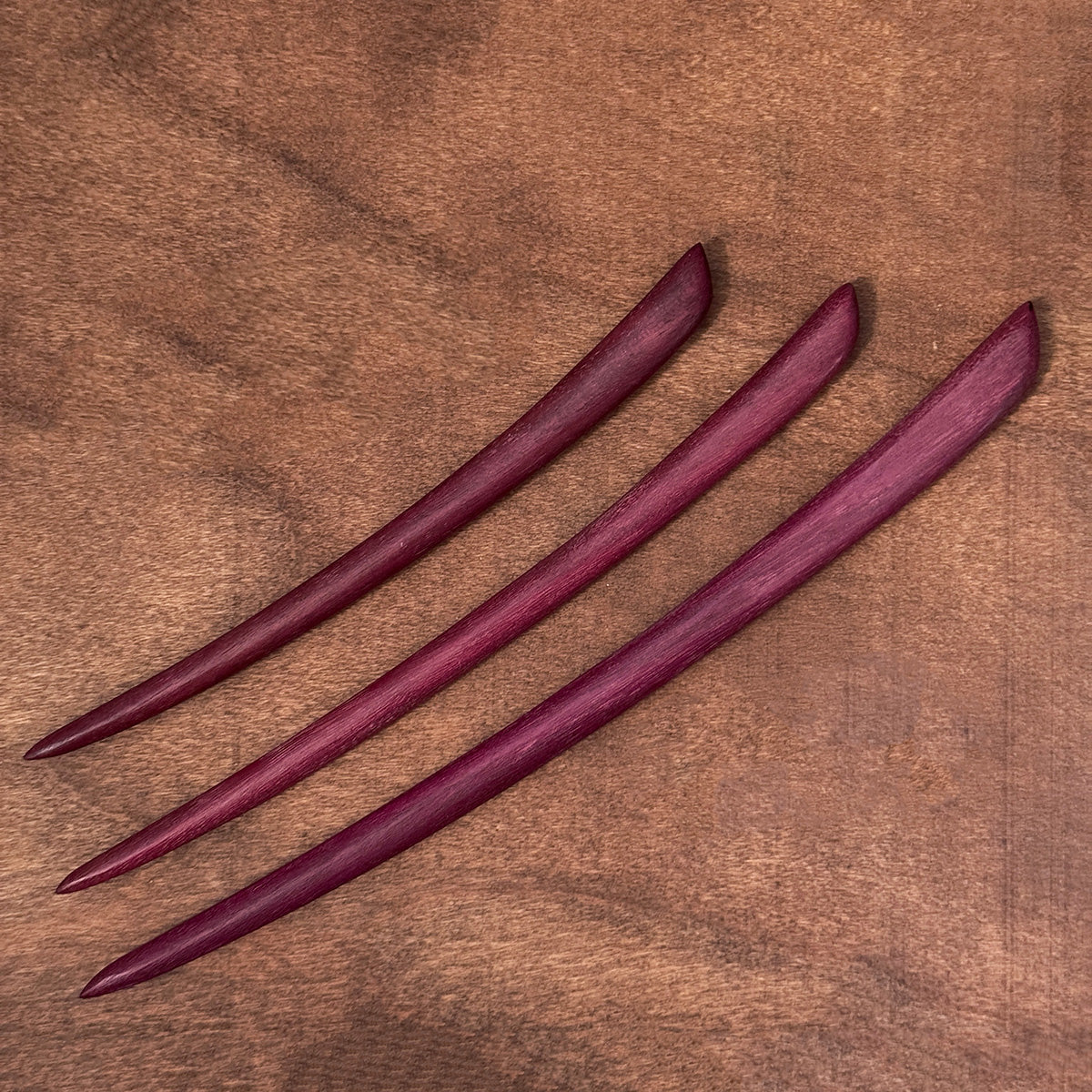 Three handmade natural purpleheart wood hair sticks arranged diagonally on a rich brown wood background.