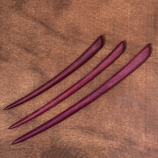 Three handmade natural purpleheart wood hair sticks arranged diagonally on a rich brown wood background.