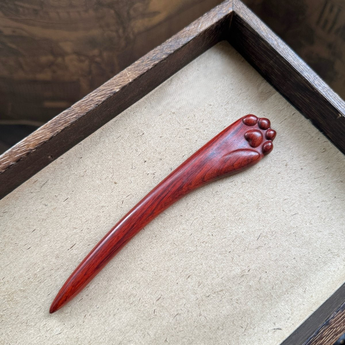 Natural rosewood hair stick with cat paw design in wooden tray on textured background.