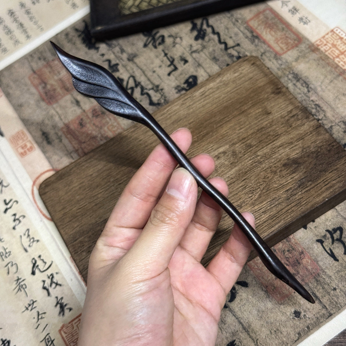 A hand holding the 18cm hand-carved ebony feather hairpin to show its detail and scale against a background of vintage paper.