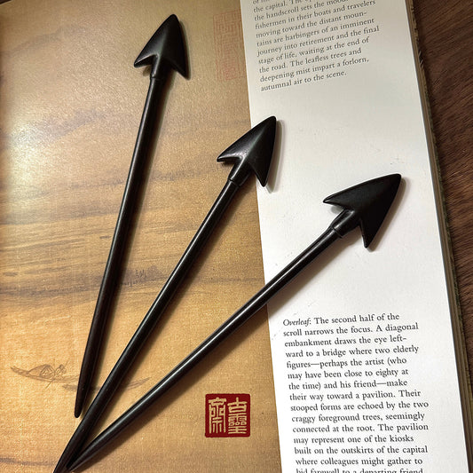 Three handcrafted Purple Sandalwood Arrow hair sticks displayed on an open art book, showing the arrow-shaped head and fine dark wood grain.