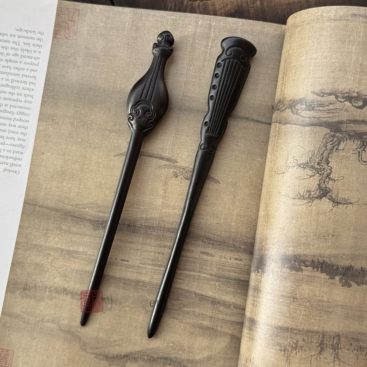 Set of two natural African Blackwood hair sticks with pipa and guqin designs on an open ancient book background.