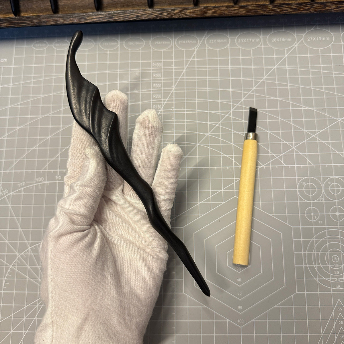 A gloved hand holding the 19.5cm ebony "Wind" hair stick next to a carving tool on a craft mat, showing its handmade quality.