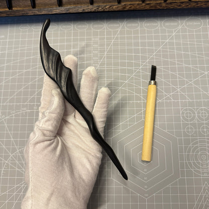 A gloved hand holding the 19.5cm ebony "Wind" hair stick next to a carving tool on a craft mat, showing its handmade quality.