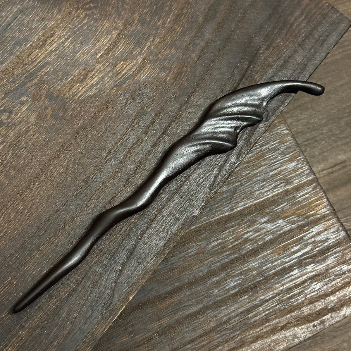 The hand-carved Violet Sandalwood "Wind" hairpin displayed on a dark, rustic wood background to highlight its texture.