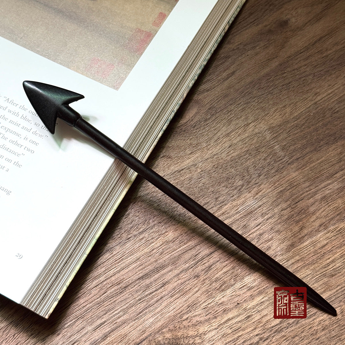 Single premium wooden Arrow hair stick made of natural Purple Sandalwood resting on a book, highlighting its sleek design and dark color for DIY crafts and updos.