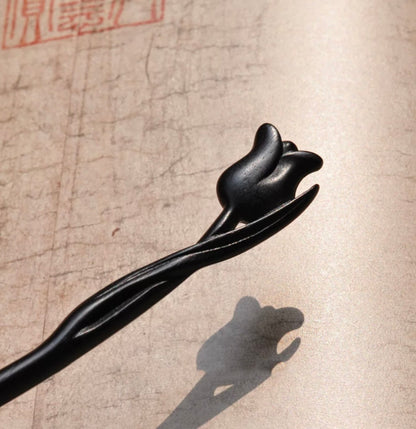 A close-up detail shot of the carved tulip head on the ebony wood hairpin, with its shadow cast on textured paper.