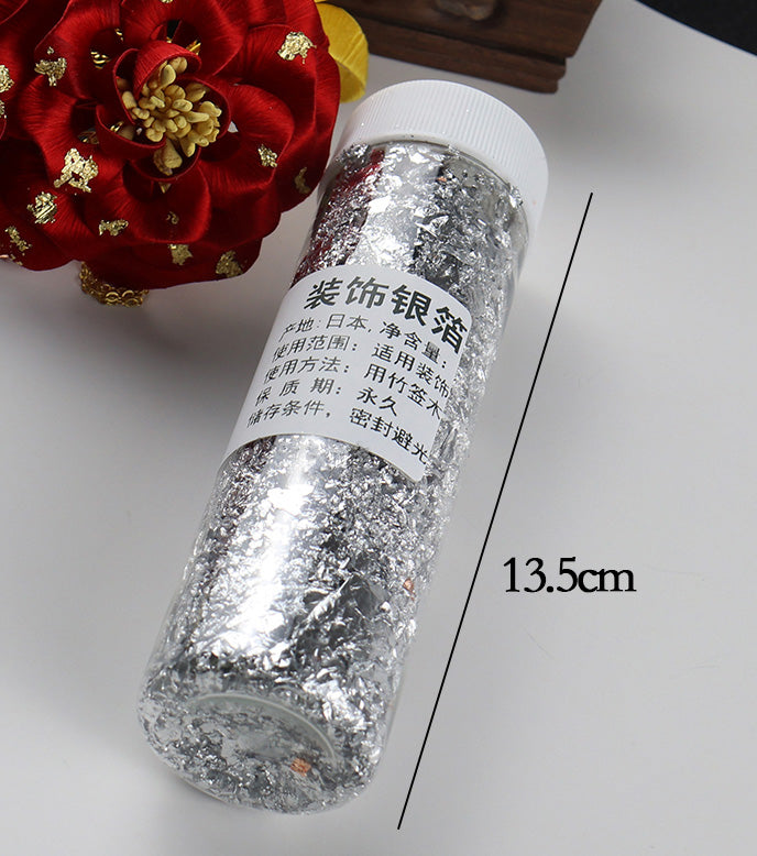 Close-up of a 13.5cm jar of decorative silver leaf flakes used for crafting and fine art.