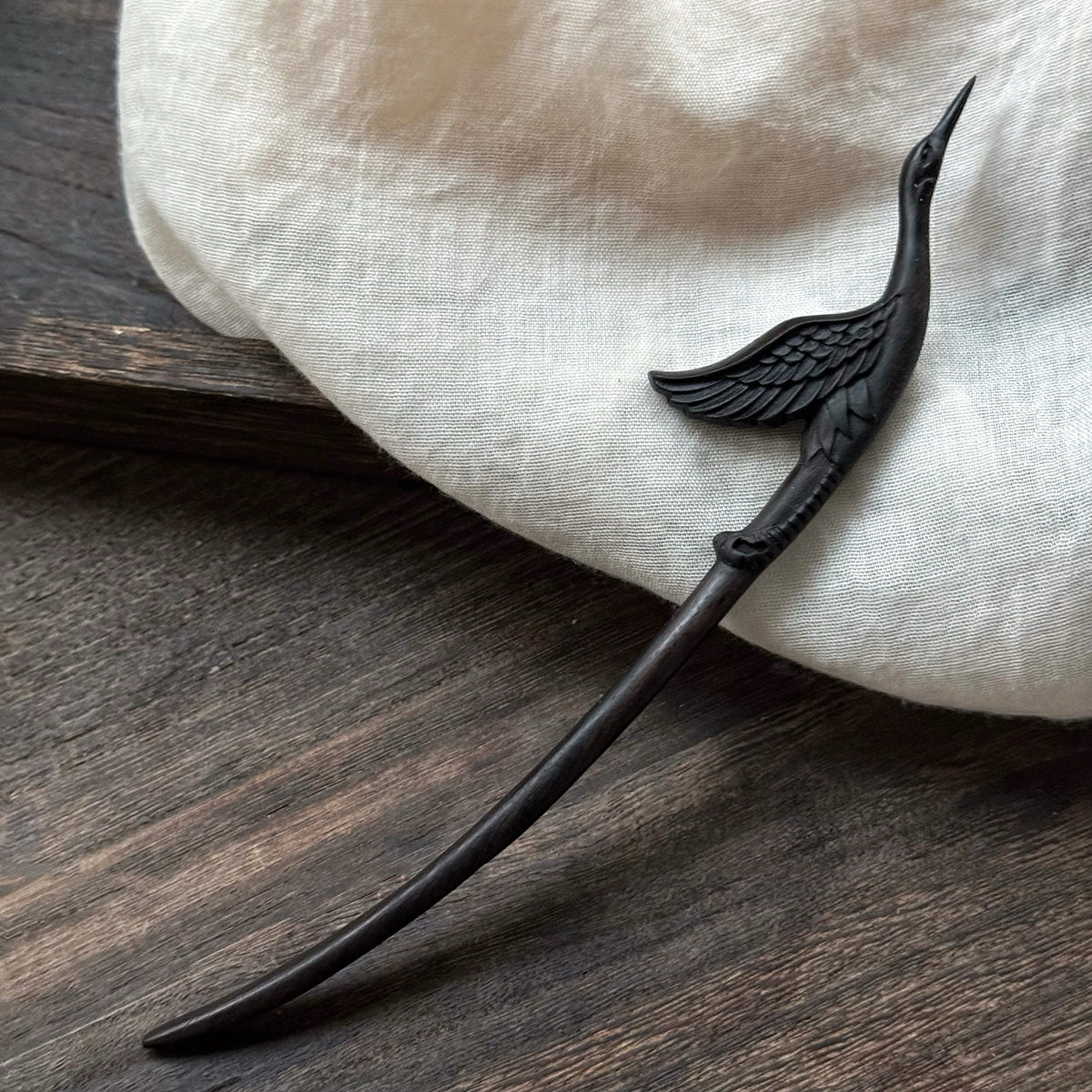Elegant black ebony wood crane hairpin resting on a white linen cloth over a dark wood surface.
