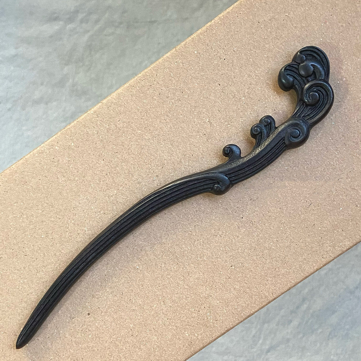 A hand-carved "Wave" design hair stick made from dark, natural African Blackwood, resting on a cardboard surface.