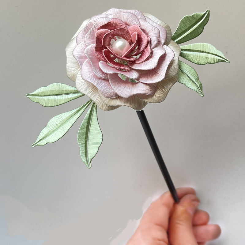 A handcrafted Yanzhi Chanhua hairpin with delicate pink and cream silk petals, a pearl center, and green leaves, held by hand against a plain background.