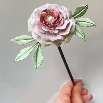 A handcrafted Yanzhi Chanhua hairpin with delicate pink and cream silk petals, a pearl center, and green leaves, held by hand against a plain background.