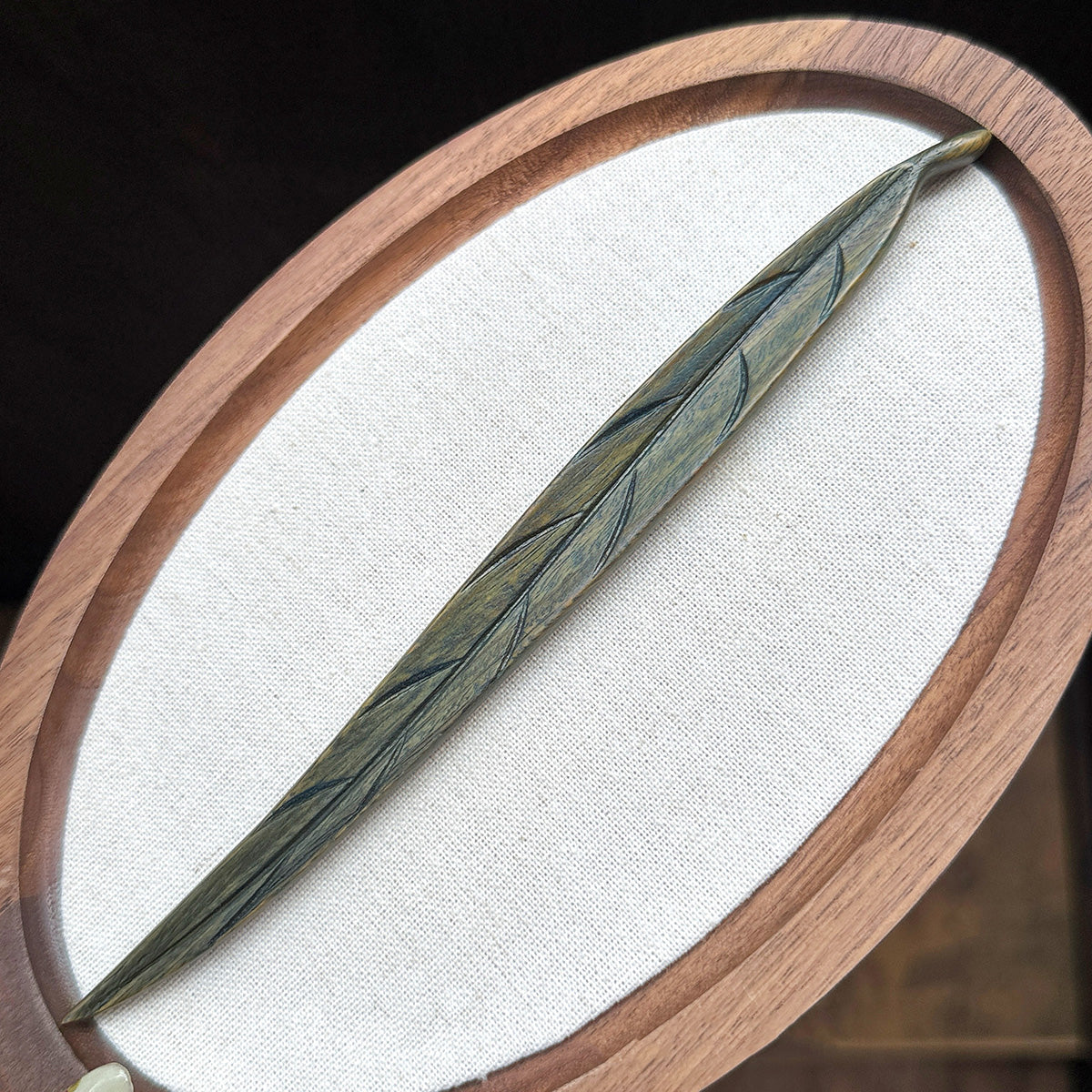A hand-carved leaf hair stick made from green sandalwood, displayed on a white oval surface with a wooden frame.