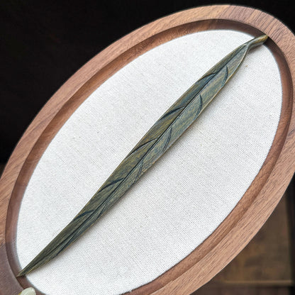 A hand-carved leaf hair stick made from green sandalwood, displayed on a white oval surface with a wooden frame.