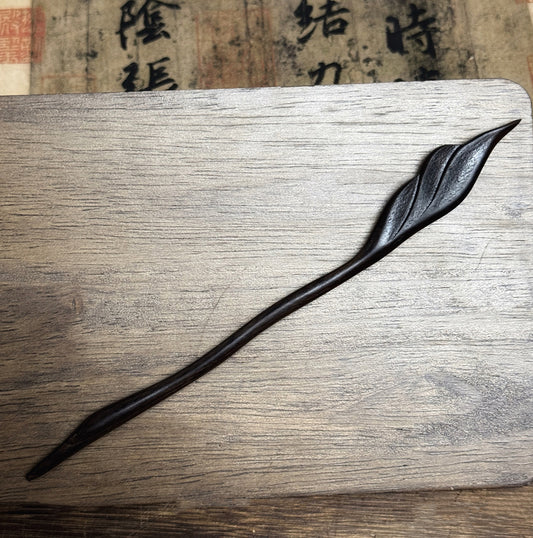 A hand-carved violet sandalwood ebony hair stick in a feather design, resting on a wooden surface with calligraphy paper in the background.