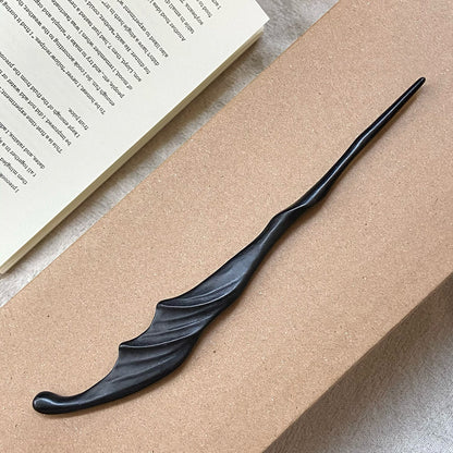 A hand-carved ebony "Wind" hair stick with a fluid, wavy design, resting on the corner of a book and a cardboard surface.