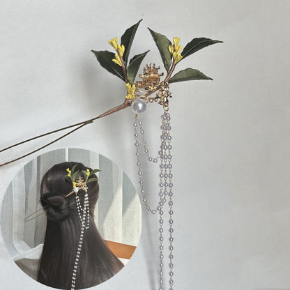 A finished handmade green and yellow fabric flower hairpin with long pearl chains, displayed as a hair accessory on a person with dark hair, highlighting the completed craft project.