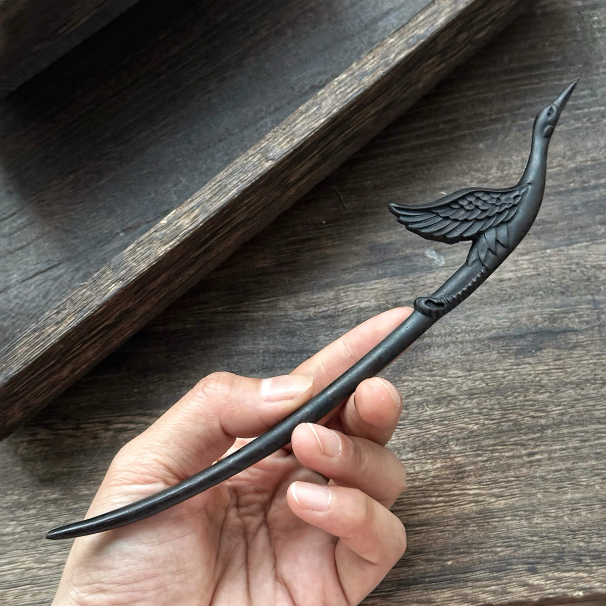 A hand holding the detailed carving of the crane on the 18cm black ebony wood hair stick to show scale.