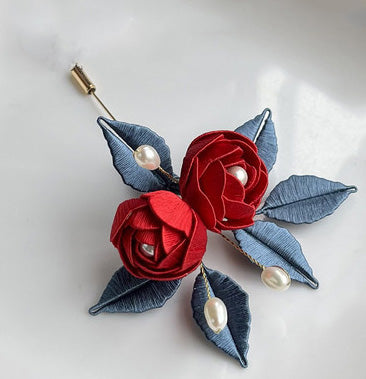 A finished handmade Chanhua rose brooch with two red silk roses, blue leaves, and pearl details on a white background.