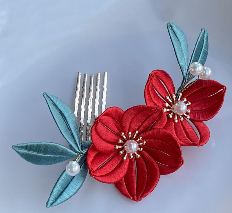 A finished handmade Chanhua hairpin with two vibrant red silk flowers and blue-green leaves attached to a silver hair comb.