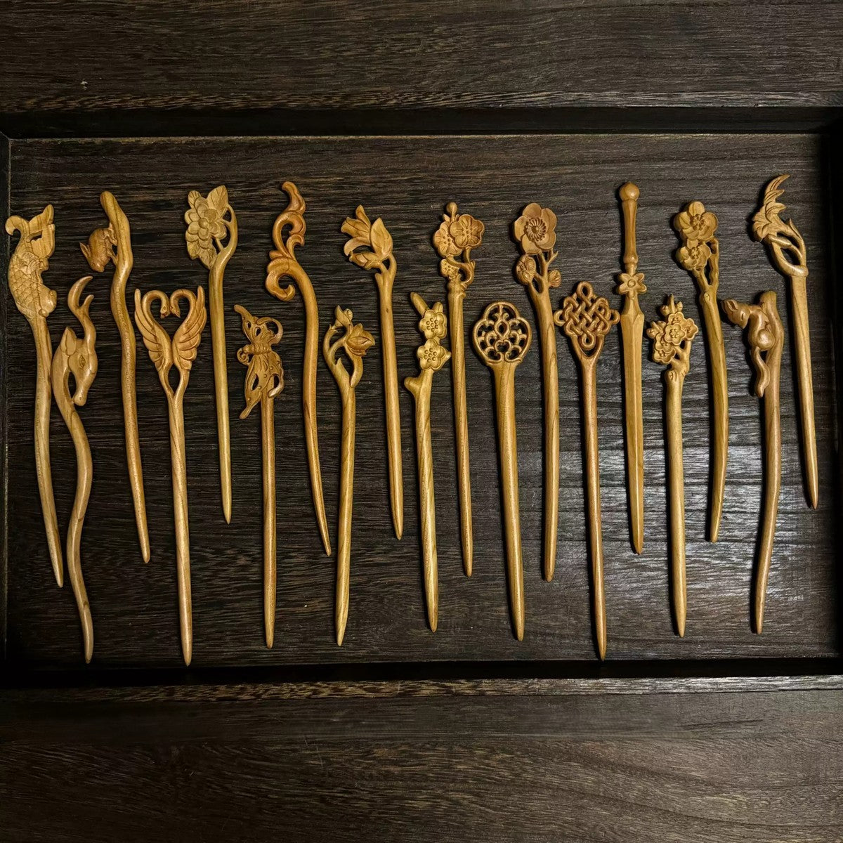 Set of natural peach wood hair sticks with assorted floral, bird, and nature carvings displayed in wooden tray