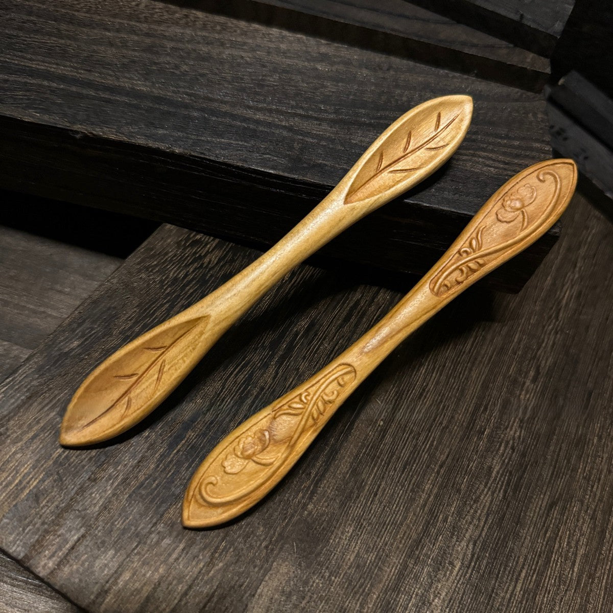 Two natural peach wood hair sticks with double-sided floral and leaf carvings displayed on dark wooden surface