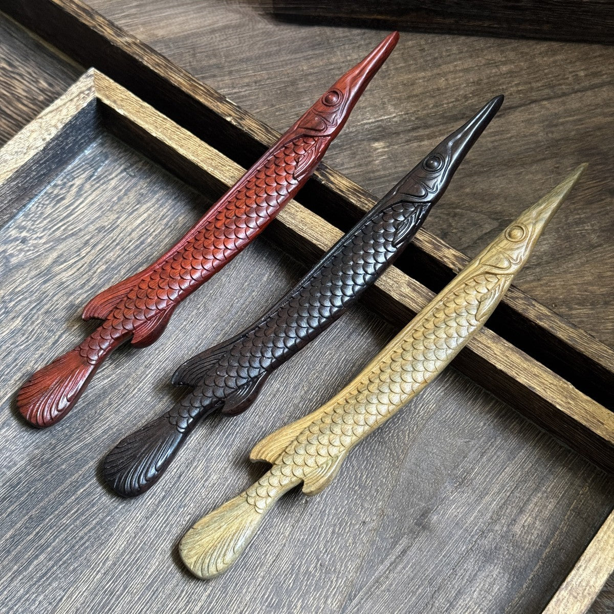 Three natural African blackwood hair sticks in fish shape design displayed in wooden tray showing color variations