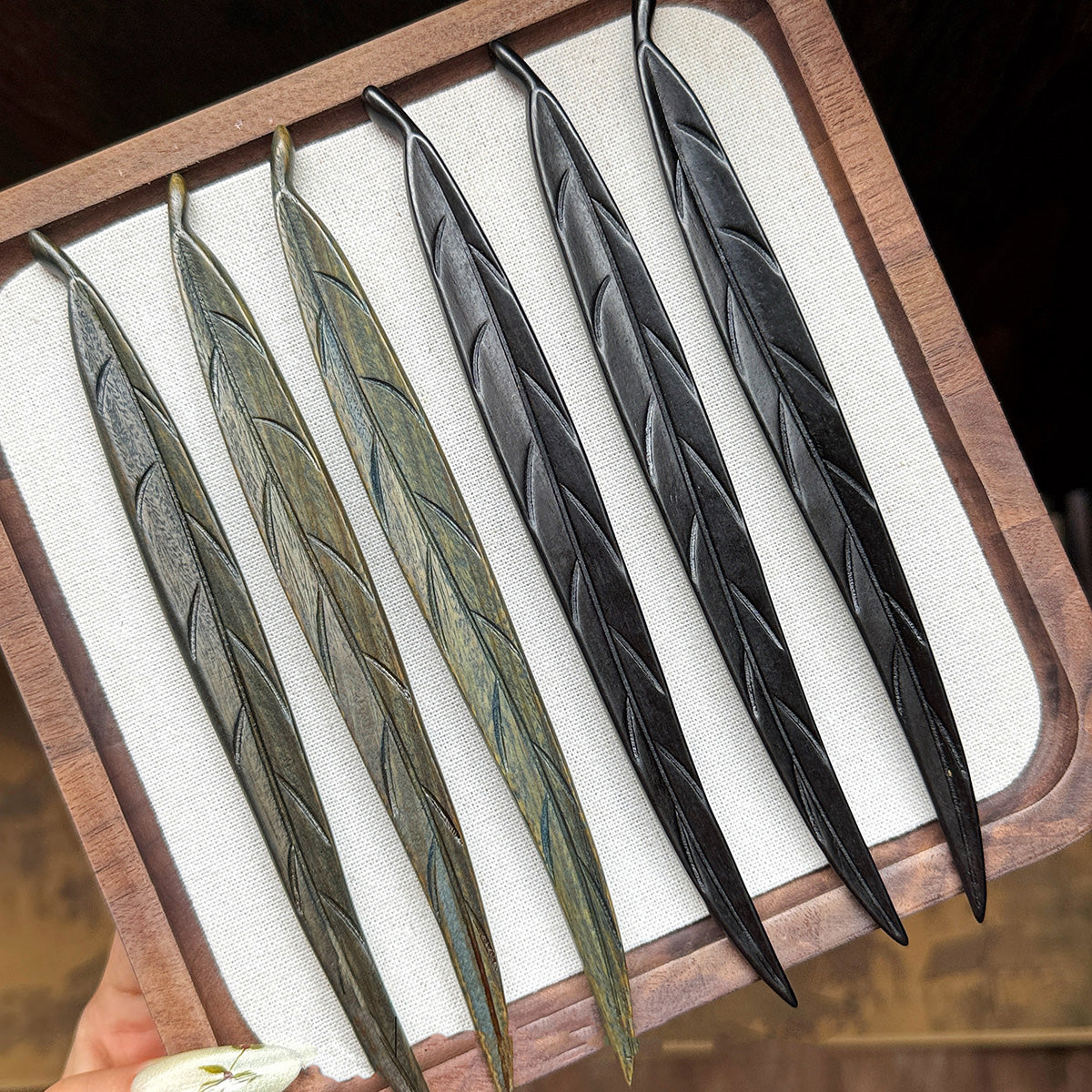 A collection of hand-carved wooden leaf hair sticks, showing the color variations between the green sandalwood and the dark ebony versions.