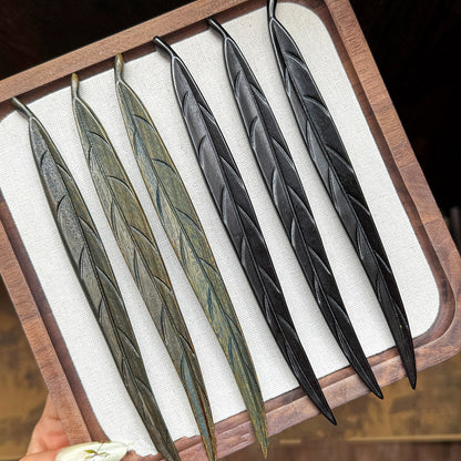 A collection of hand-carved wooden leaf hair sticks, showing the color variations between the green sandalwood and the dark ebony versions.