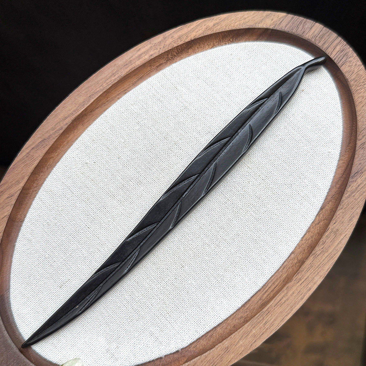 A hand-carved leaf hair stick made from dark ebony wood (violet sandalwood), displayed on a white oval surface with a wooden frame.
