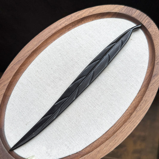 A hand-carved leaf hair stick made from dark ebony wood (violet sandalwood), displayed on a white oval surface with a wooden frame.