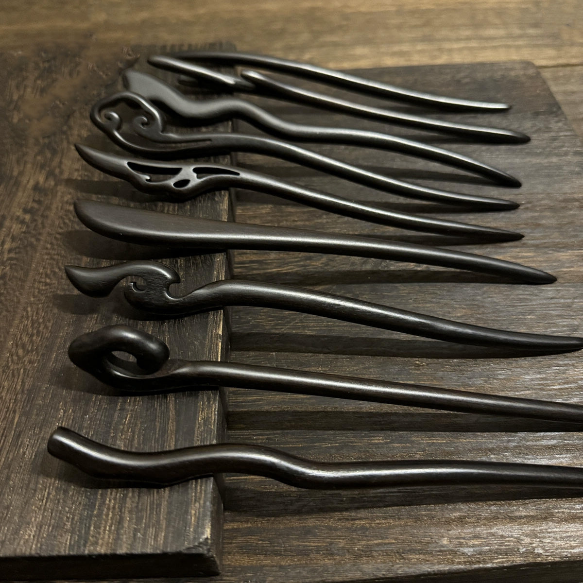 A collection of unique handmade black sandalwood hair sticks, showcasing various carved designs, laid out on a dark wood board.
