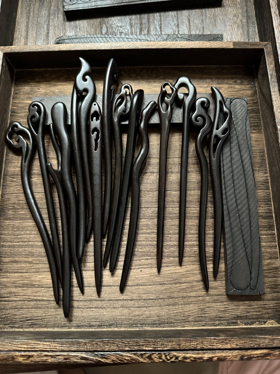 A collection of assorted hand-carved black sandalwood hair sticks displayed in a rustic wooden tray, highlighting their unique shapes and smooth finish.