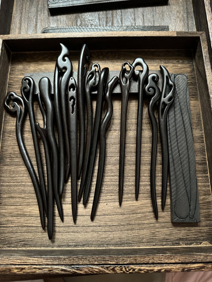 A collection of assorted hand-carved black sandalwood hair sticks displayed in a rustic wooden tray, highlighting their unique shapes and smooth finish.