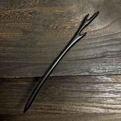 A hand-carved natural black sandalwood hair stick with a minimalist forked top, resting on a dark wood background.