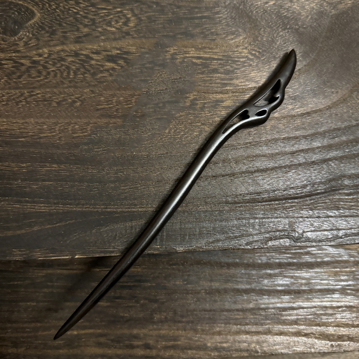 An intricately carved black sandalwood hair pin with a flowing, wing-like design at the top, shown on a textured wooden surface.