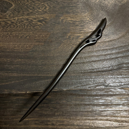 An intricately carved black sandalwood hair pin with a flowing, wing-like design at the top, shown on a textured wooden surface.