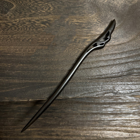 An intricately carved black sandalwood hair pin with a flowing, wing-like design at the top, shown on a textured wooden surface.