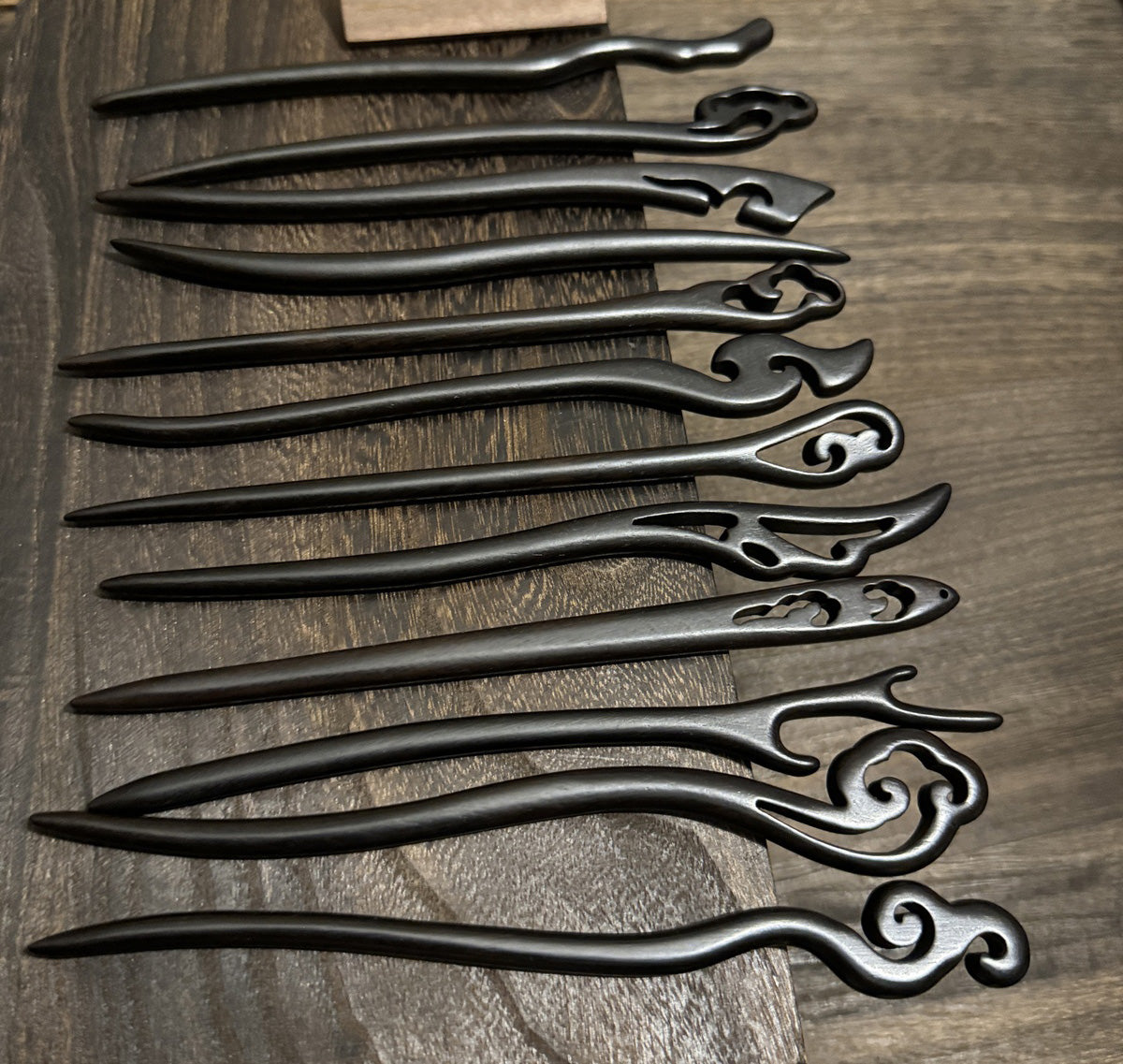 A collection of 12 unique, hand-carved black sandalwood hair sticks with intricate cloud and scroll designs, arranged horizontally on a dark wood surface.